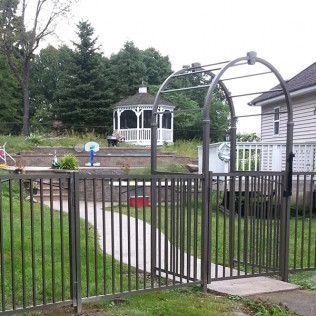 A metal gate with an archway leads to a backyard path with a gazebo and trees.