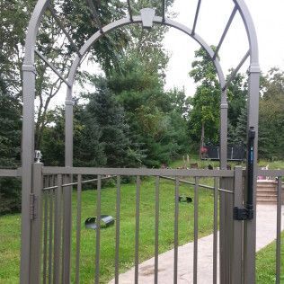 Metal garden gate with an arched top, leading to a grassy yard with trees in the background.