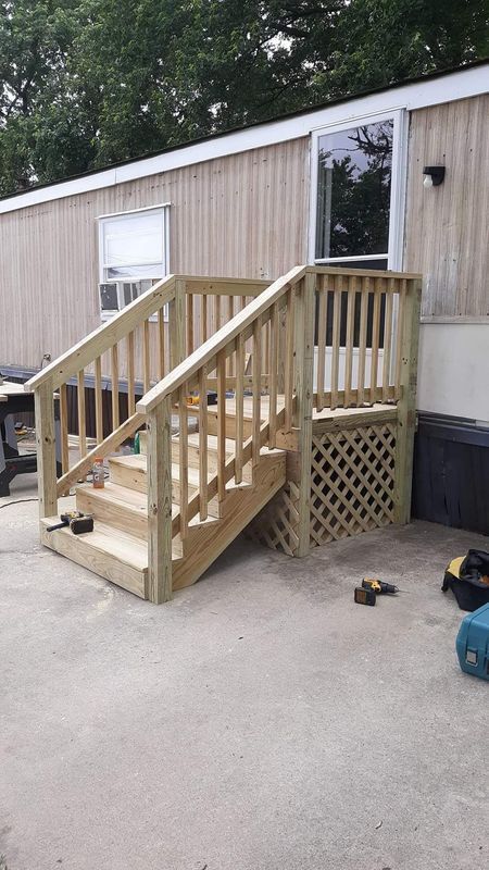 Wooden stairs leading to a mobile home's entrance. The stairs have railings, a lattice enclosure, and sit on a concrete patio.