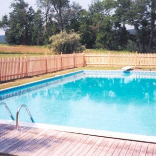 Swimming pool with wooden deck, diving board, and picket fence in a grassy yard with trees.