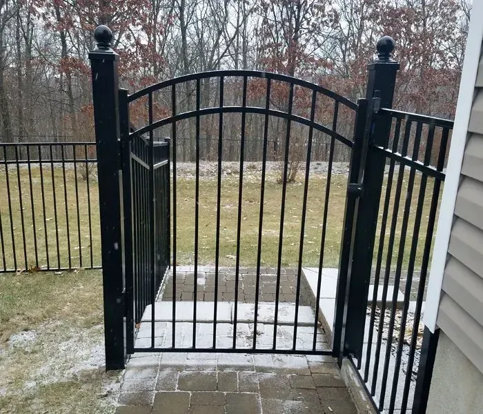 Black metal gate in a fenced backyard, opening onto a patio. Winter setting, trees in the background.