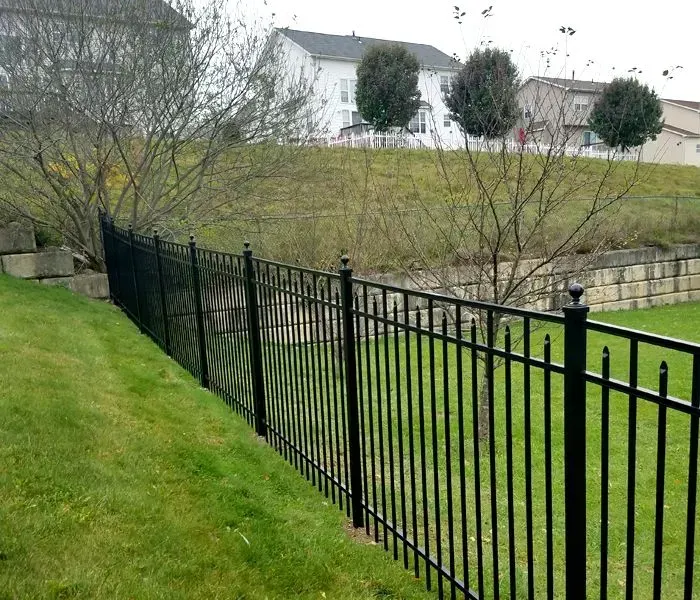 Black metal fence on green lawn, separating yard from sloped area with trees and houses.