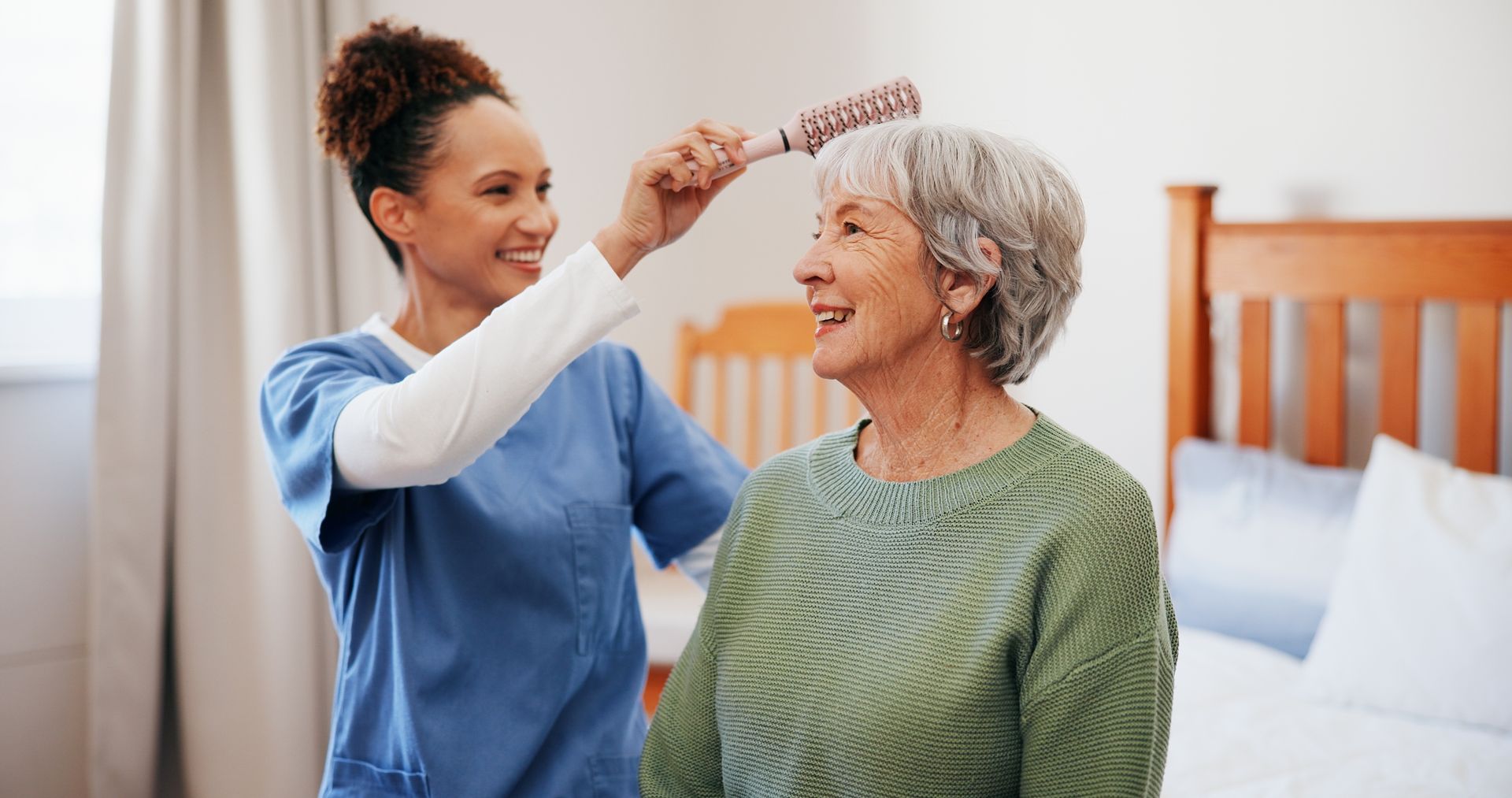Caregiver combing an older woman's hair in a bedroom, both smiling