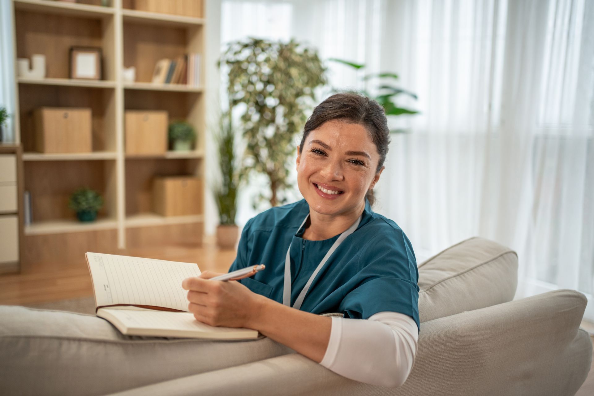 Person smiling on a sofa, holding a pen beside an open notebook in a bright living room.