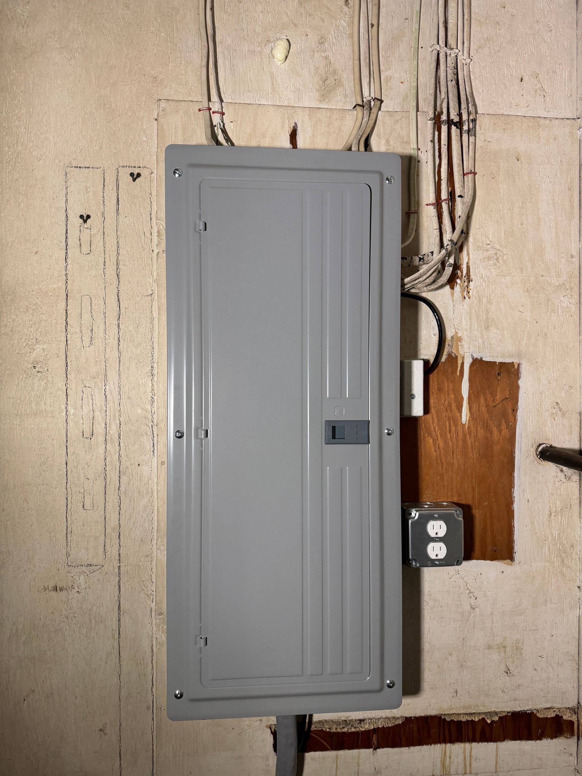 Gray electrical panel mounted on a concrete wall. Wires and outlet nearby.