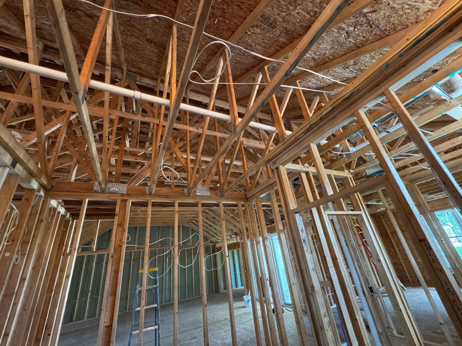 Interior of a house under construction. Wooden framework of walls and roof visible, with electrical wiring and pipes.