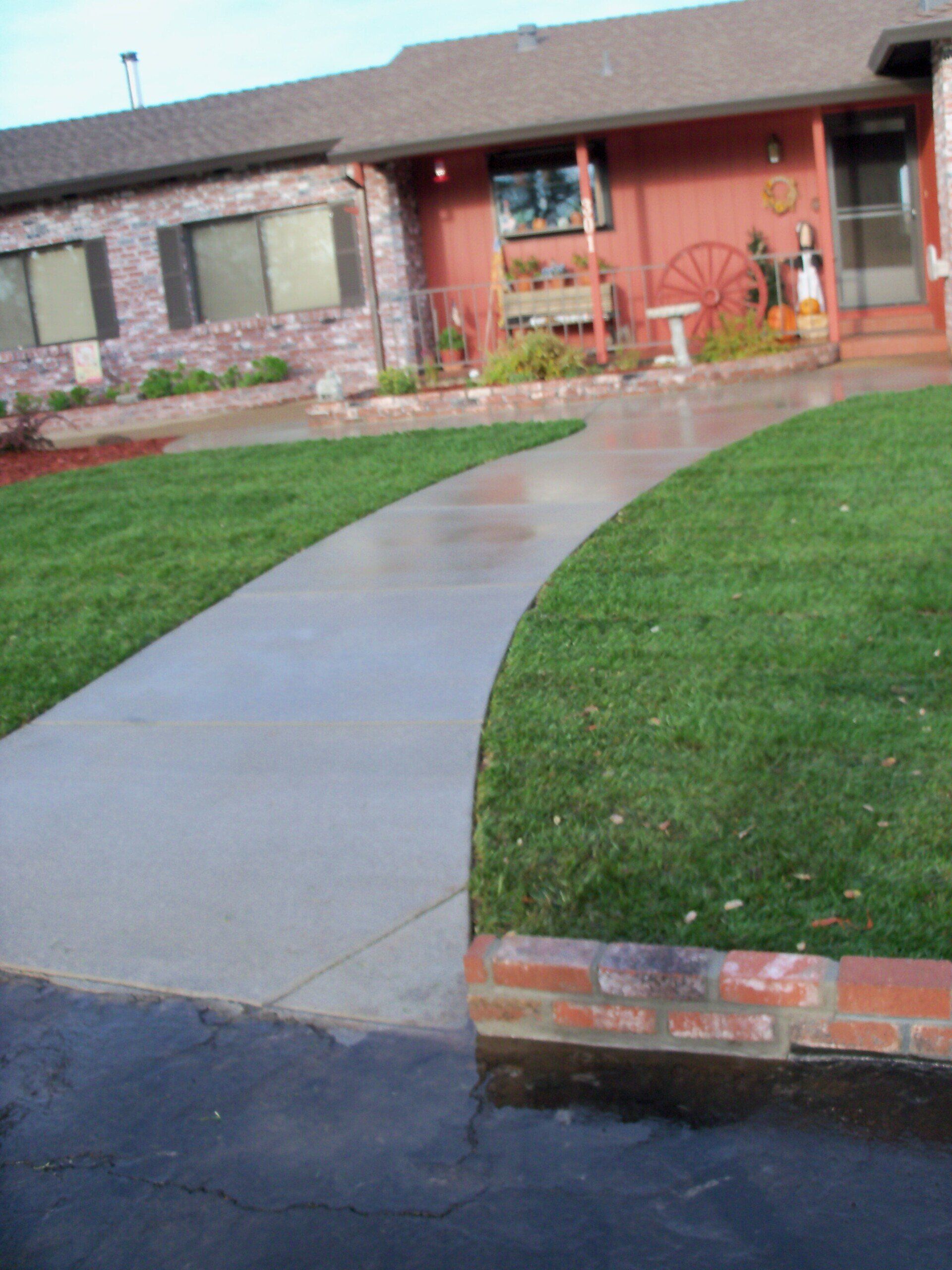 Concrete walkway curving from driveway to a reddish-brown house with green lawn and brick edging.