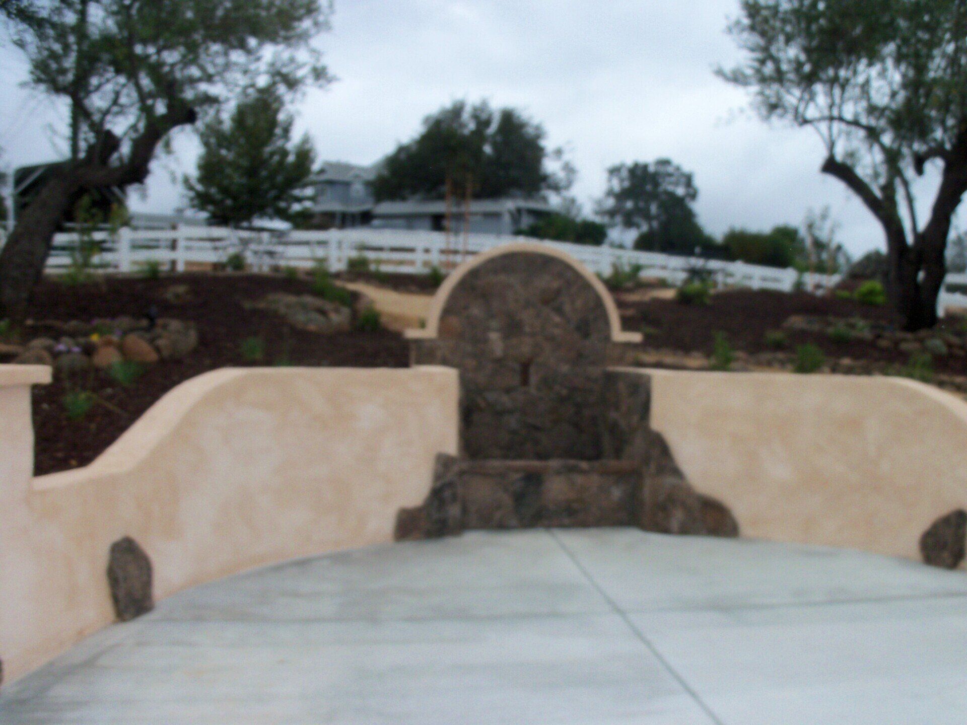 A stone water fountain built into a stucco wall, with a concrete patio in front, and trees in the background.