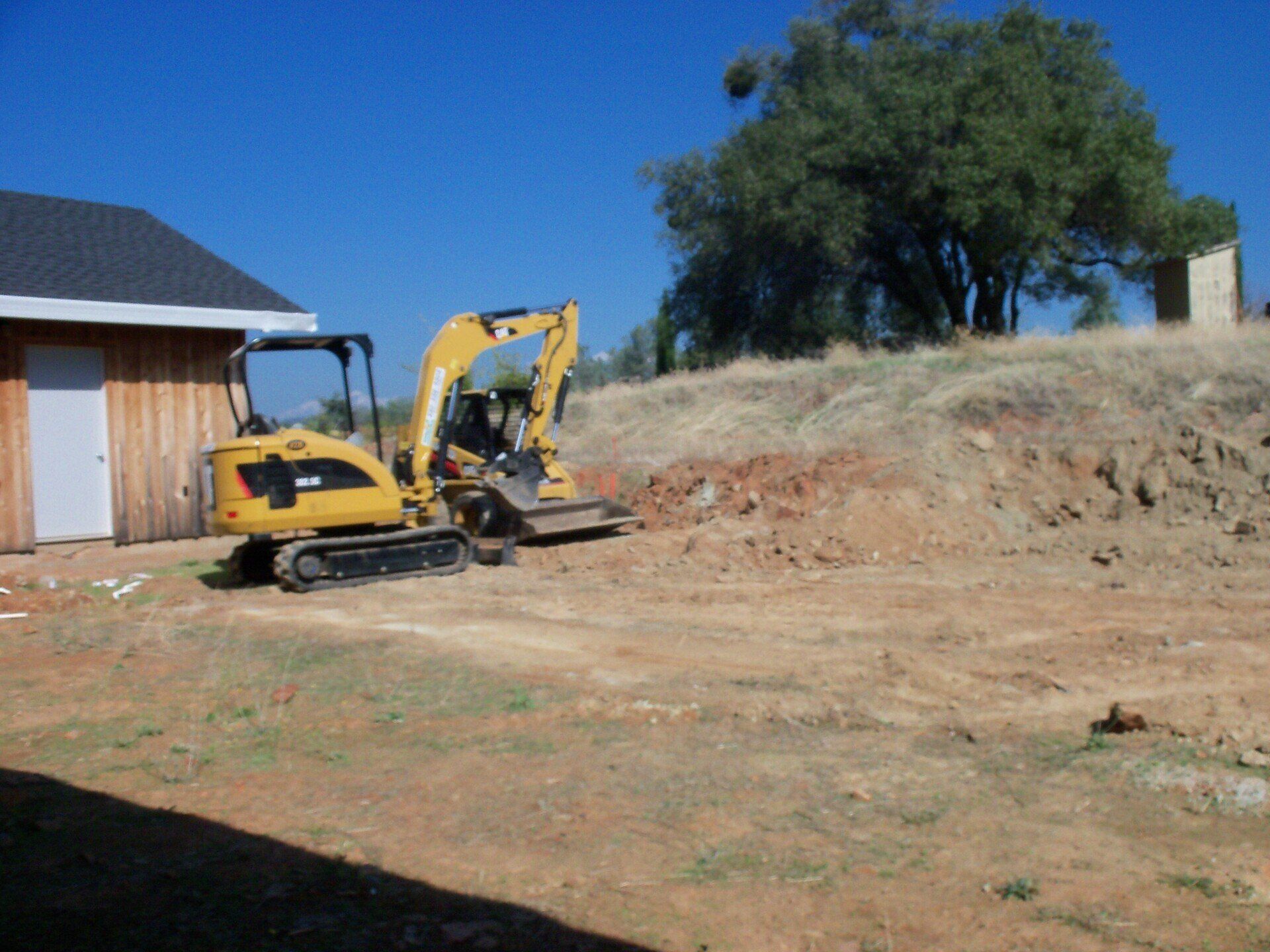 Yellow excavator digging dirt near a light brown shed on a sunny day.