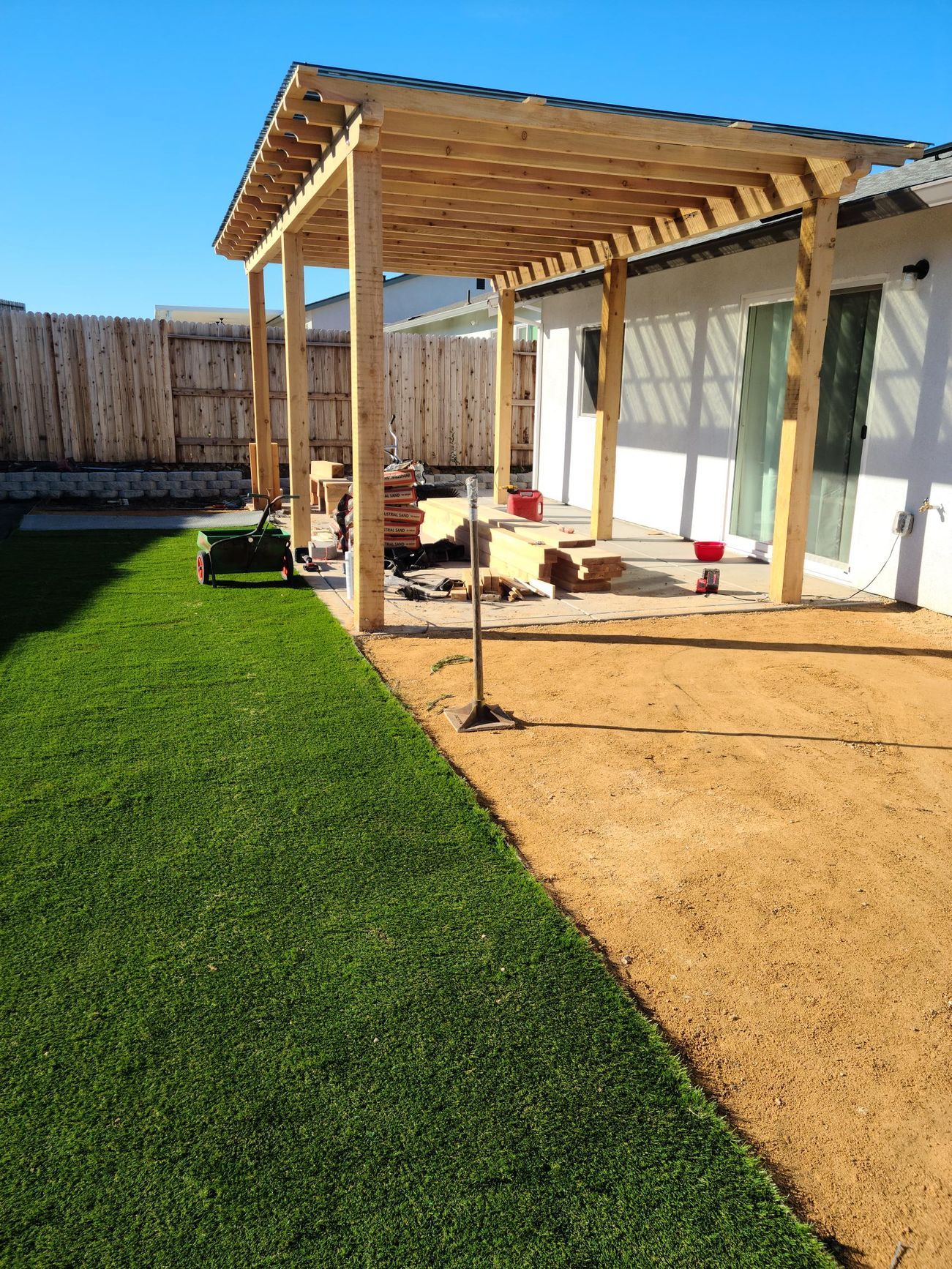 A wooden pergola attached to a house over a gravel patio and grass. Sunny outdoor setting.