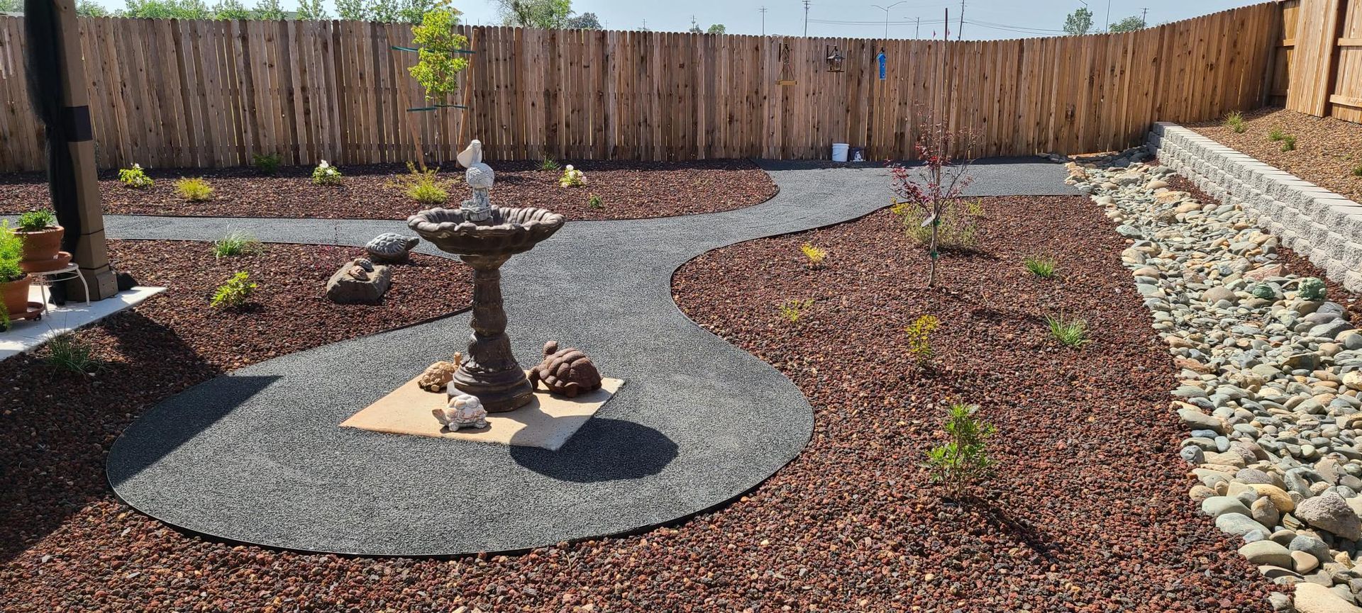 A backyard with a winding stone path, fountain, and mulch landscaping. Wooden fence in the background.