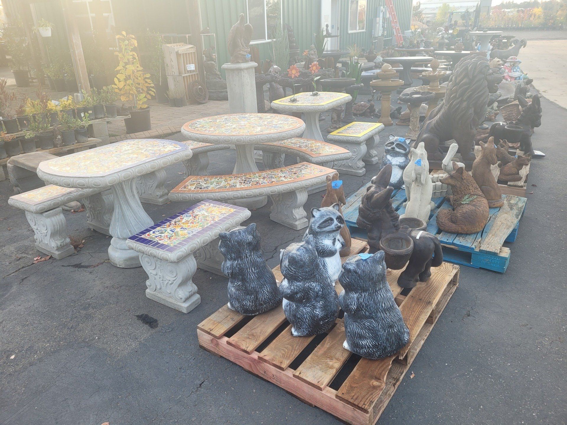 Outdoor display of garden decor, including concrete tables and benches, and raccoon statues on pallets.
