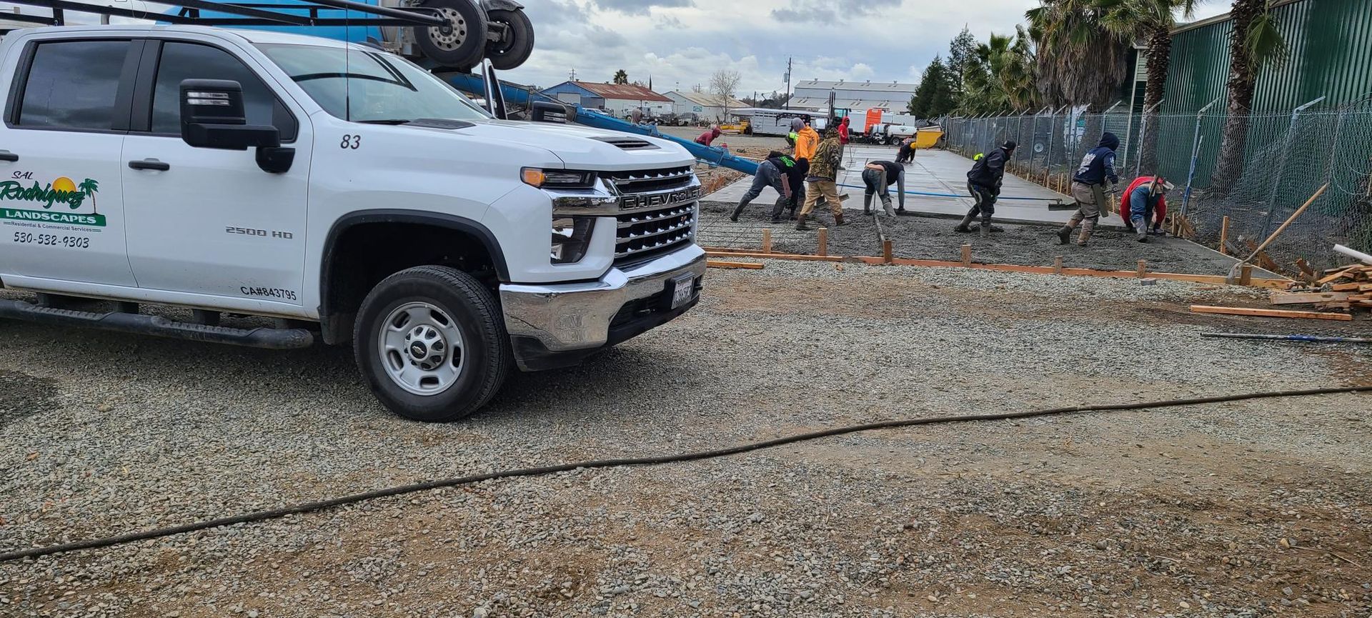 A white pickup truck parked near construction workers laying gravel, cloudy sky.