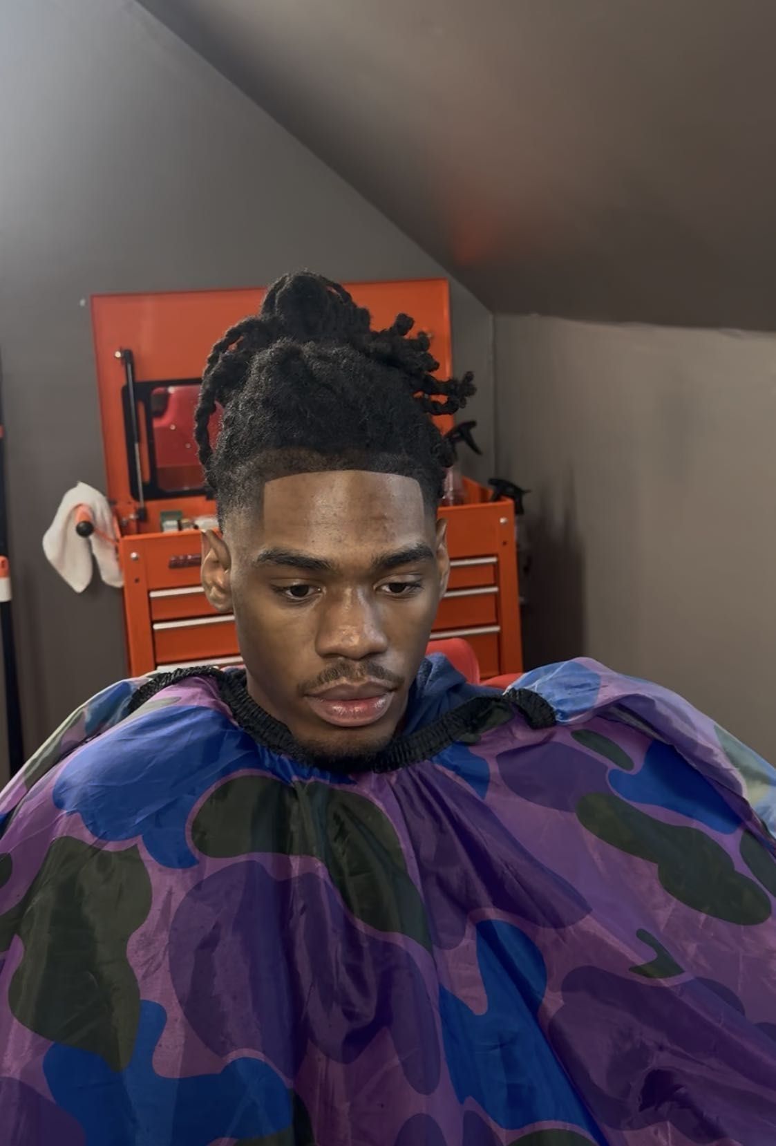 Person seated indoors with a purple camouflage barber cape, looking down in a room with an orange tool chest.