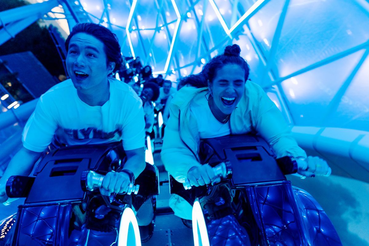 People on illuminated roller coaster, smiling and reacting excitedly. Blue lights.