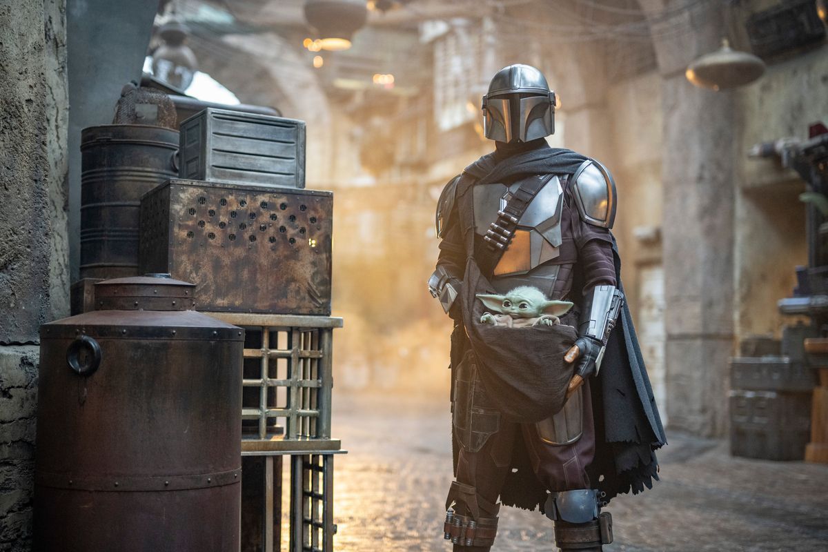 The Mandalorian with Grogu in a dusty corridor, flanked by metal containers and dimly lit hanging lamps.