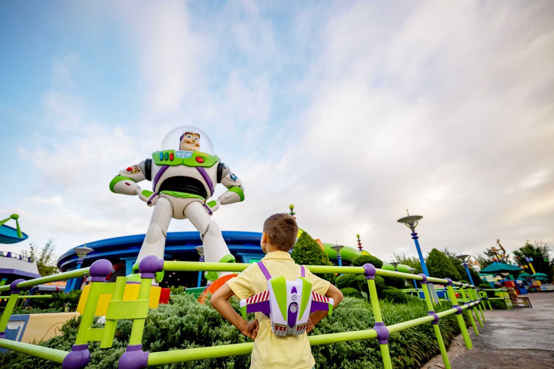 Boy with Buzz Lightyear backpack, standing before a large Buzz Lightyear statue at Toy Story Land in a park.