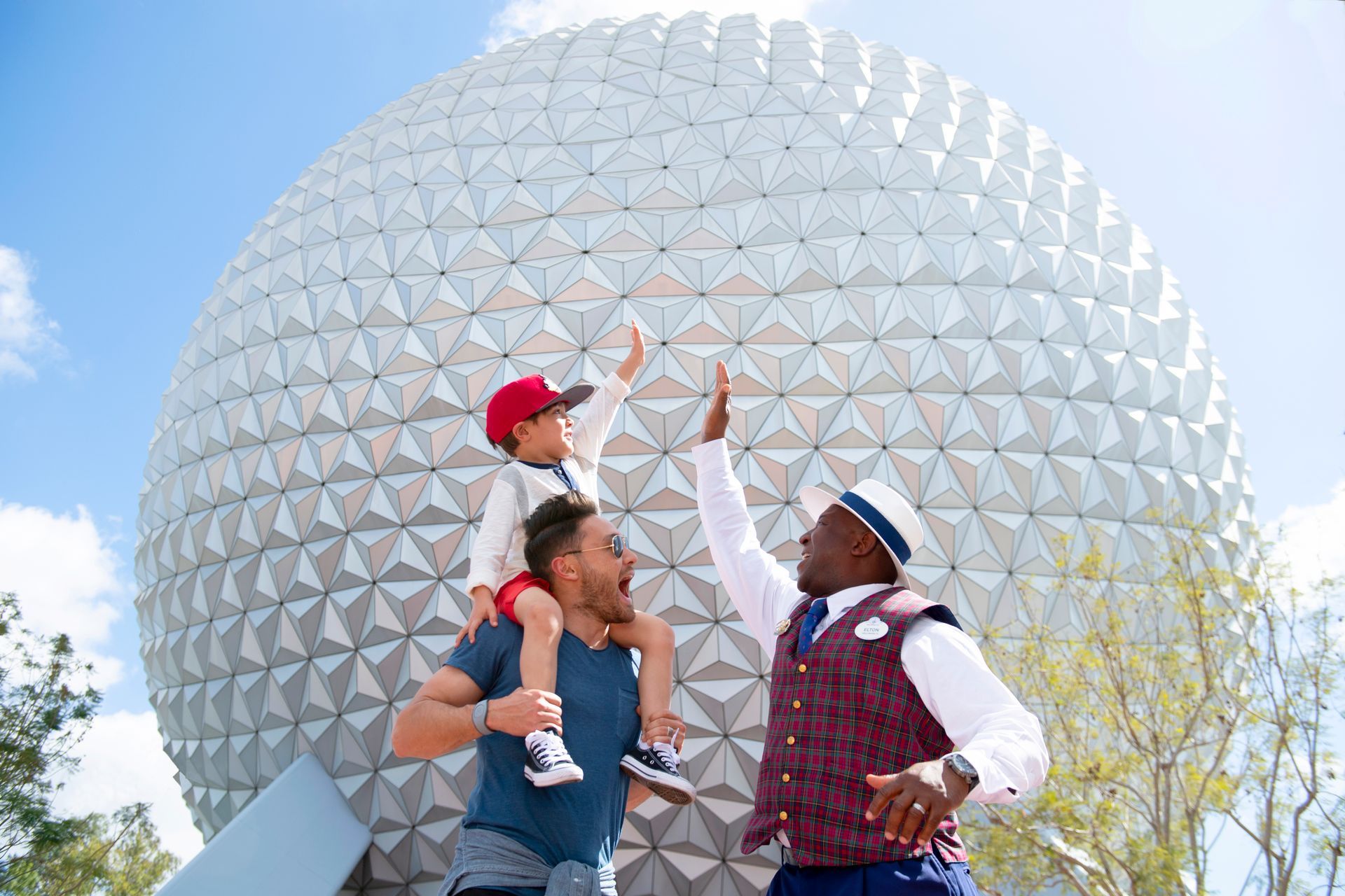 Man holding child on shoulders, high-fiving cast member in front of Spaceship Earth at Epcot.