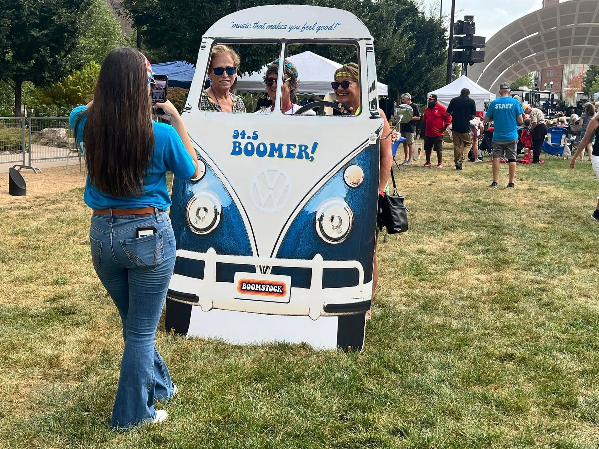 A woman is taking a picture of people standing behind a cardboard VW that says boomer.