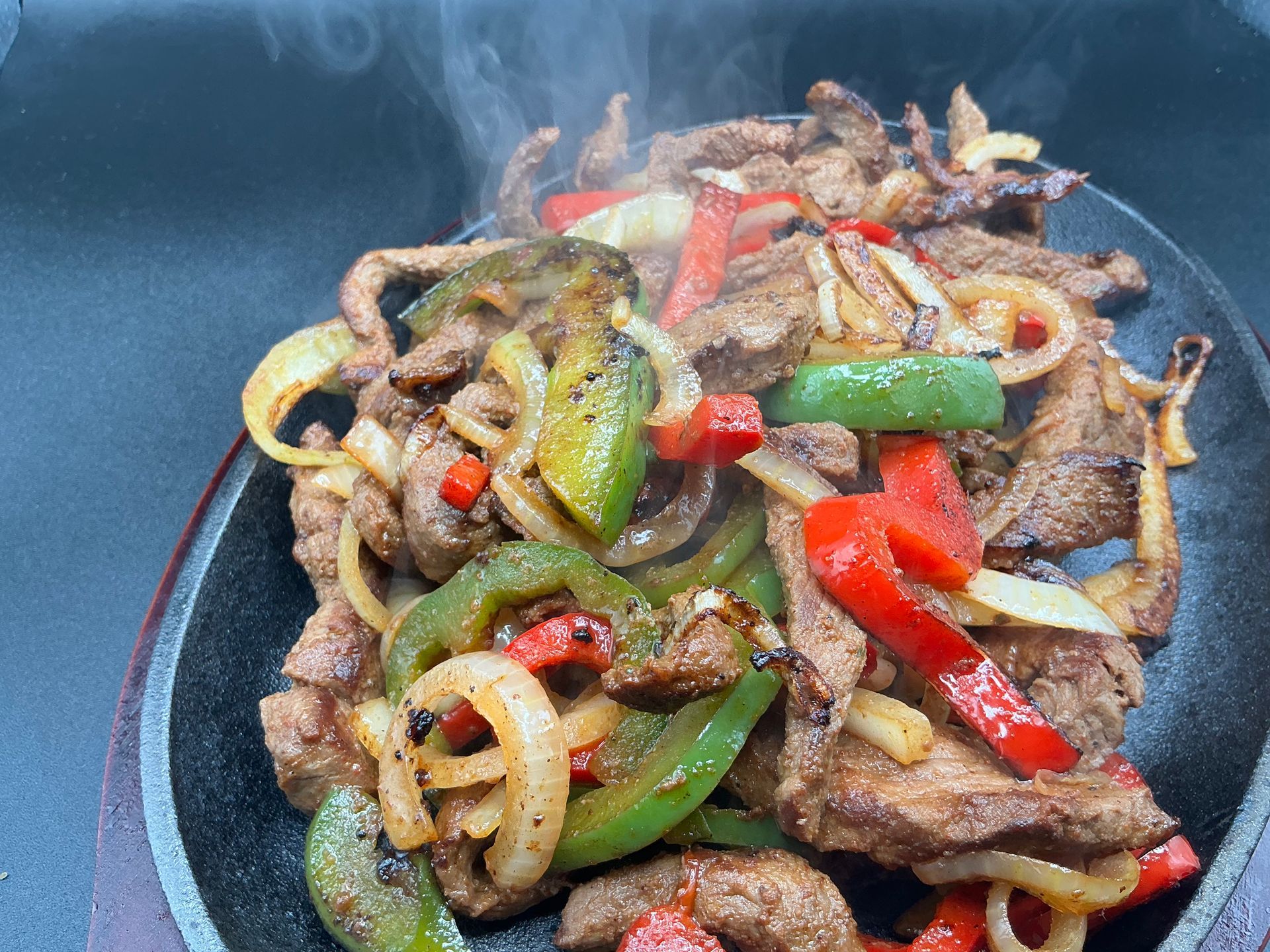Sizzling fajitas: steak strips, onions, and bell peppers on a hot, dark skillet. Steam rises.