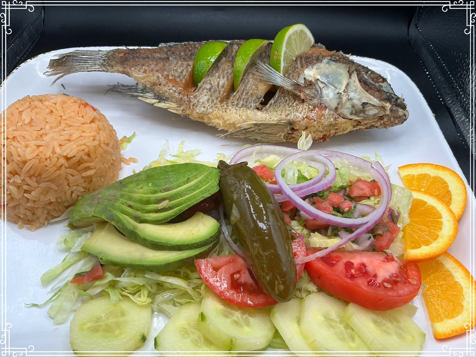 Fried fish dish with rice, avocado, and vegetables. Slices of lime and orange.