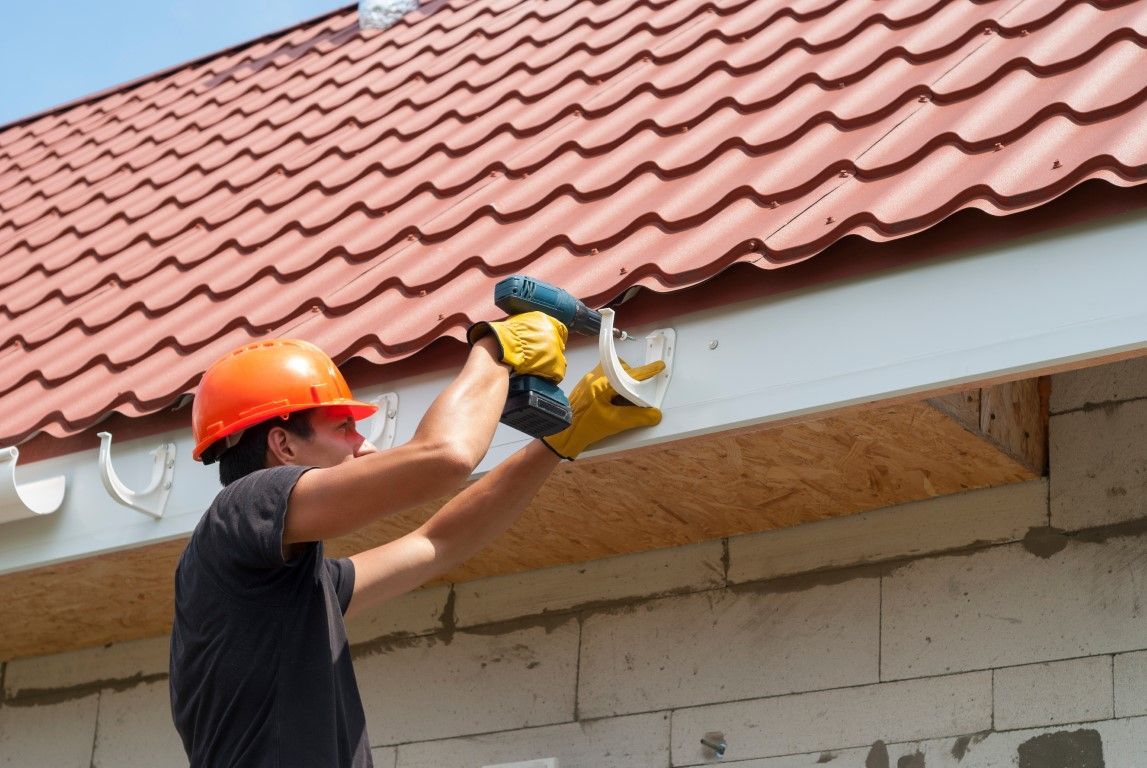Man in hard hat drills into gutter on house with red roof.