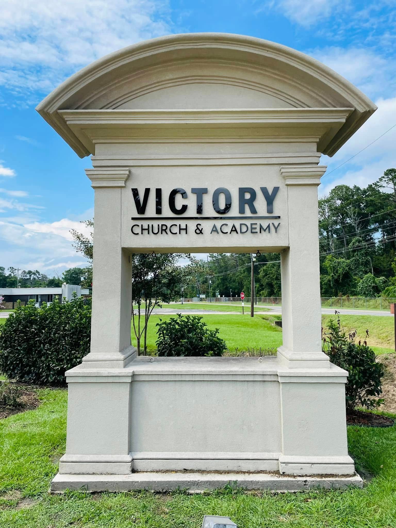 Sign for Victory Church & Academy, cream-colored structure with text, set in green grass with trees.