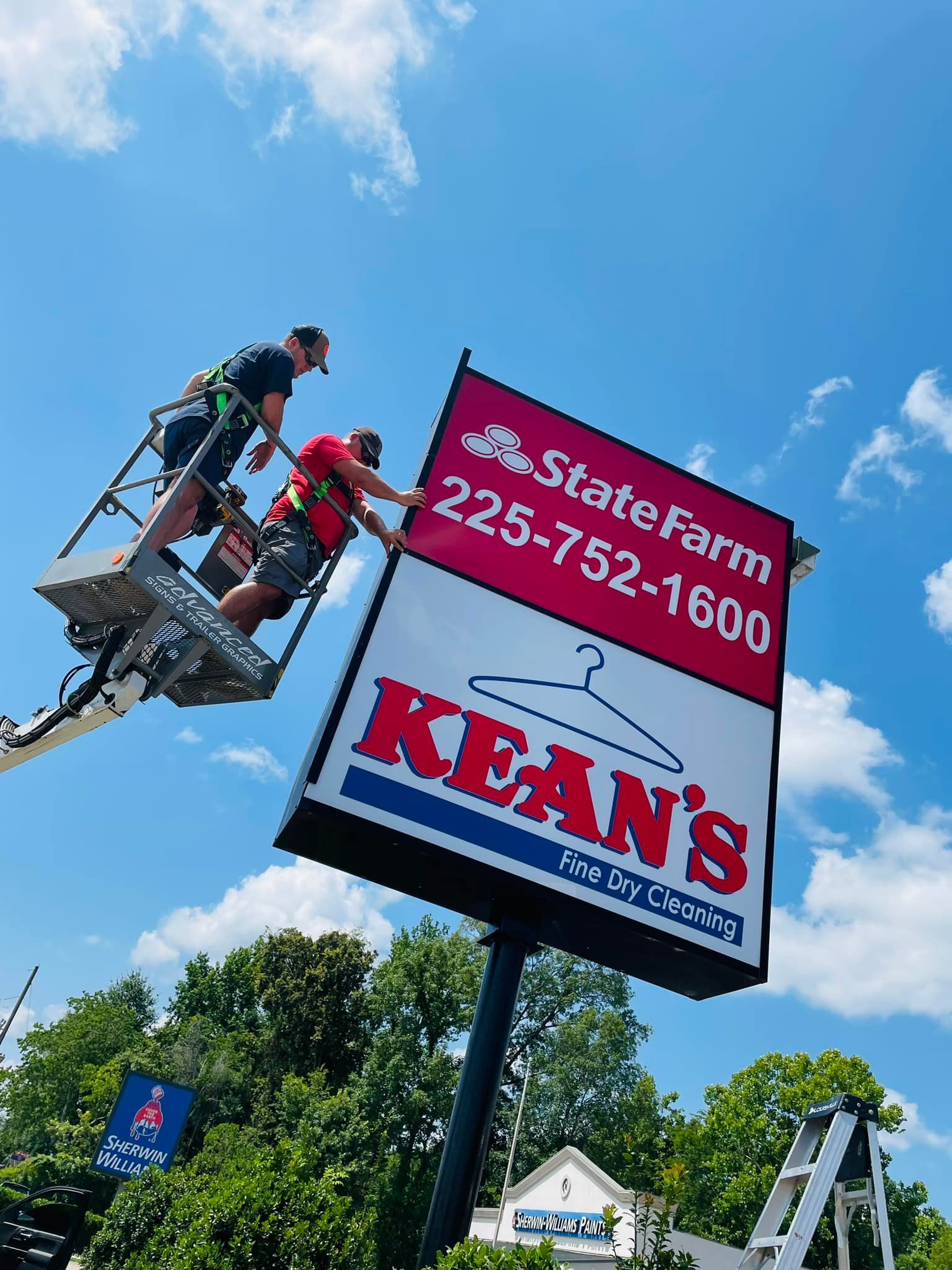 Two men on a lift installing a sign for Kean's Cleaners and State Farm against a blue sky.