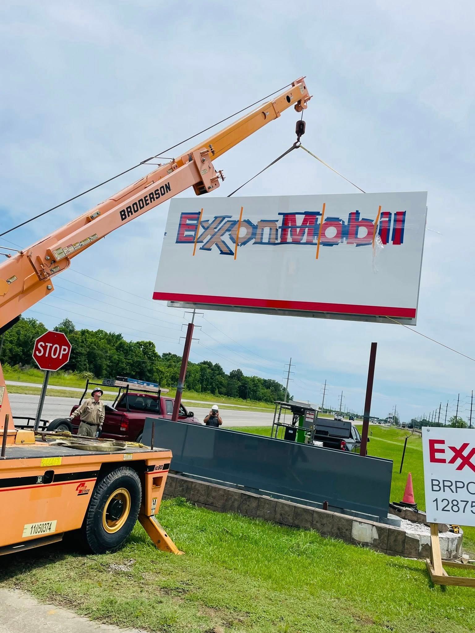 Crane installing an ExxonMobil sign, with a gray barrier in front of a grassy area under a sunny sky.