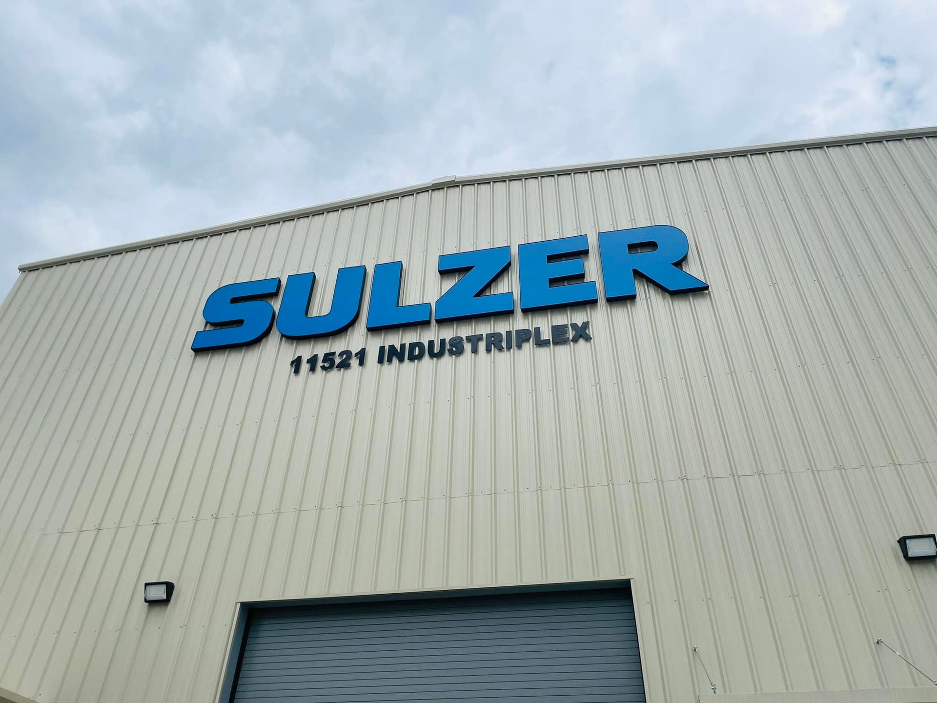 SULZER sign on a white industrial building, address 11521 Industriplex, under cloudy sky.