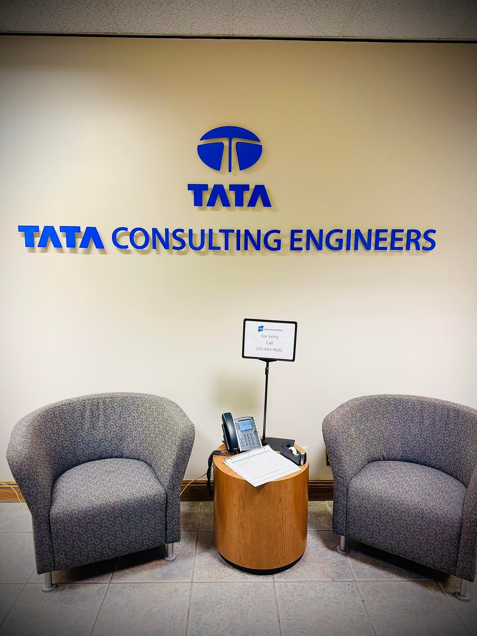 Two gray chairs face a Tata Consulting Engineers logo on a white wall. A small table with items sits between the chairs.