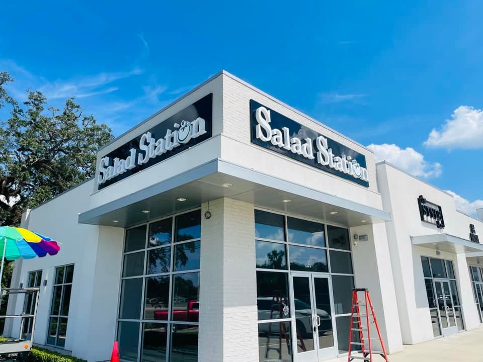 Exterior of a Salad Station restaurant with white walls and large windows under a blue sky.