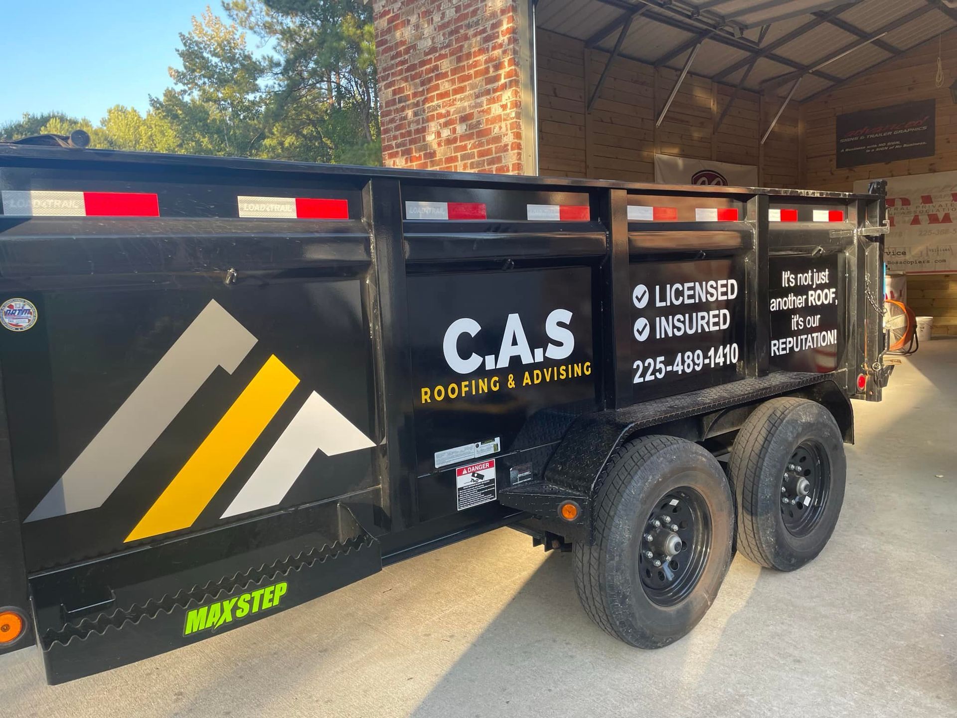 Black dump trailer with C.A.S. Roofing & Siding logo and contact info, parked outdoors.