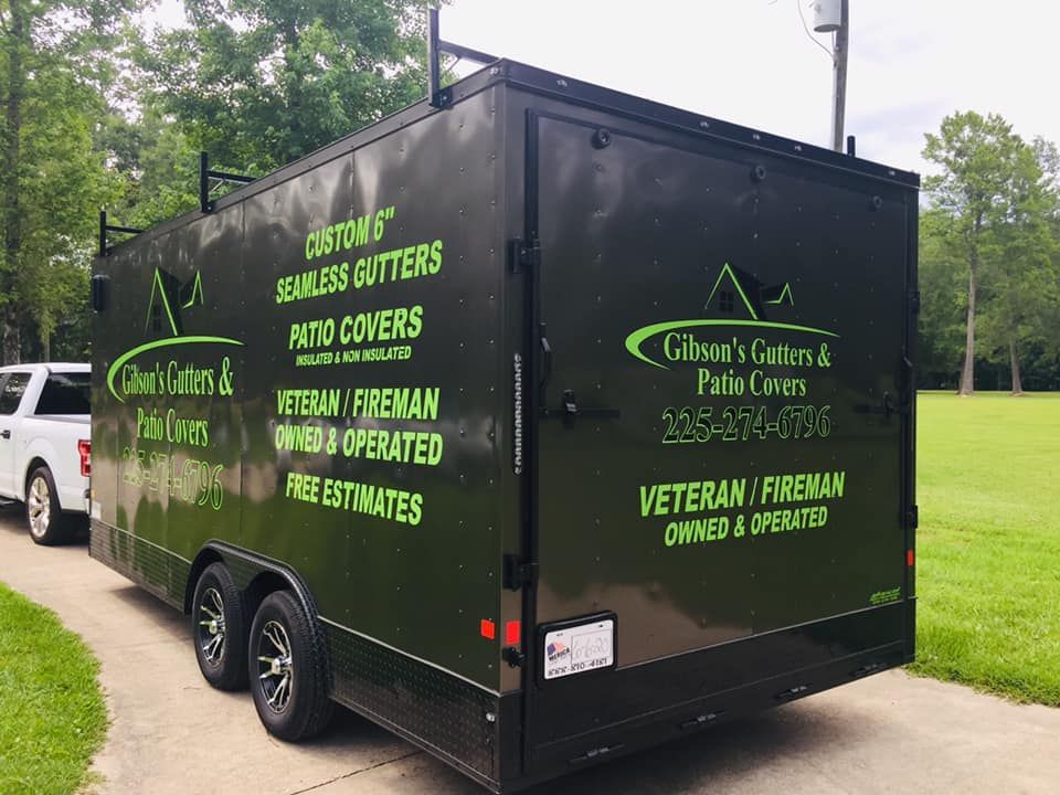 A black trailer with green text and logos for Gibson's Gutters & Patio Covers, parked on a driveway.