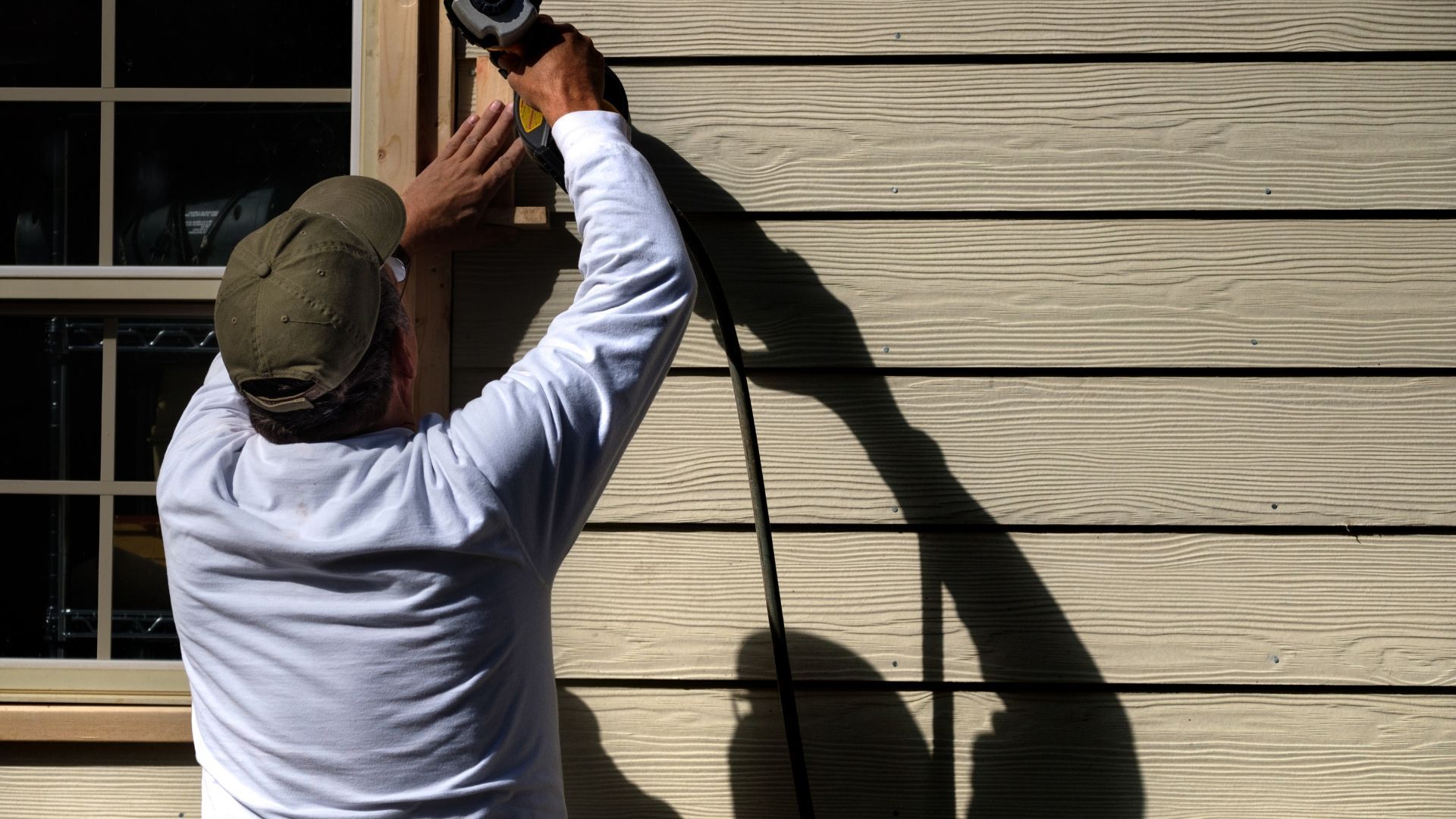 A person wearing a white long-sleeved shirt and green cap uses a nail gun to install siding next to a window.