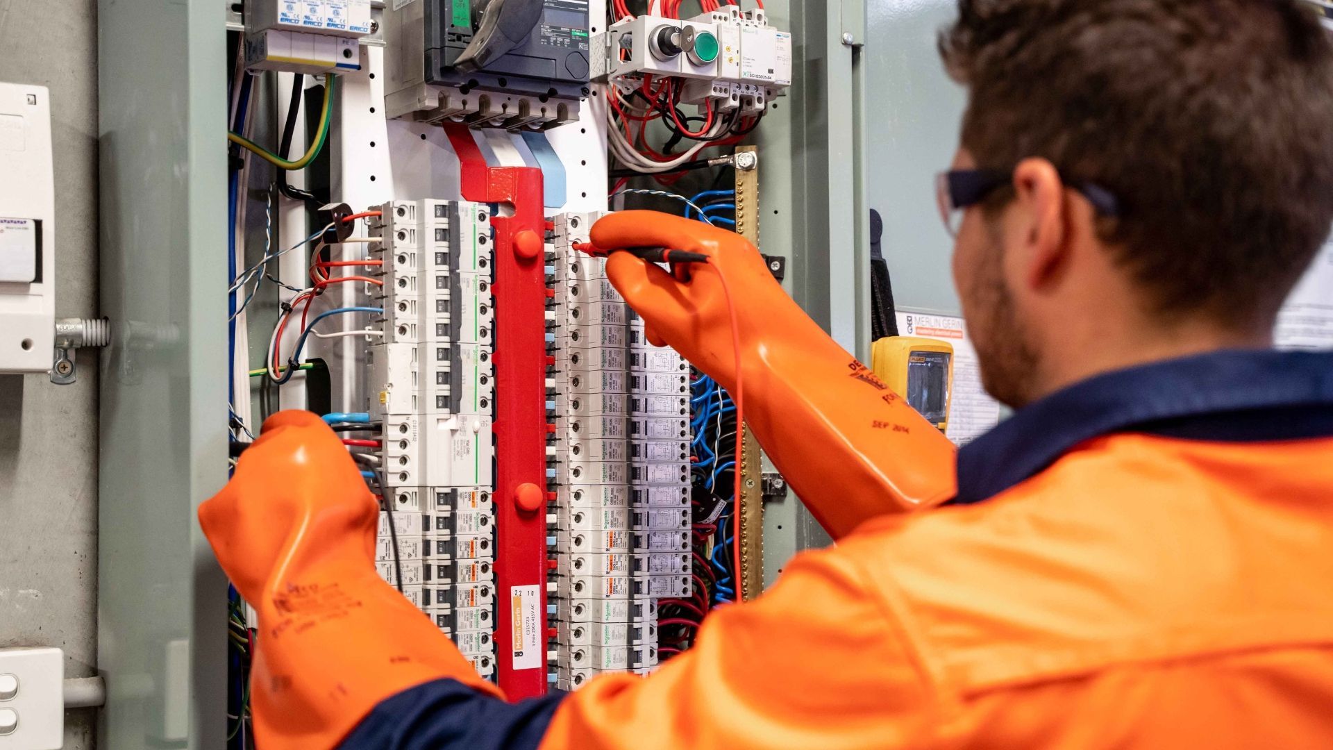 A technician in safety gear and orange insulated gloves inspects the electrical components of a wall-mounted panel.