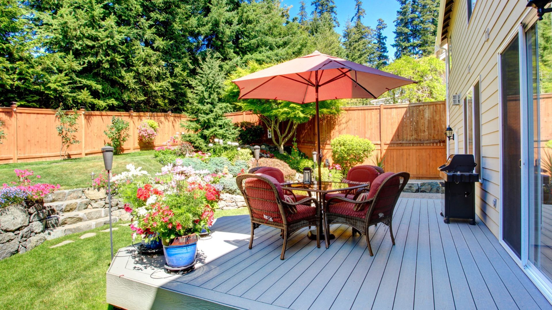 A grey deck features a red umbrella, four chairs, and a potted plant, set against a fenced backyard with a lawn and trees.