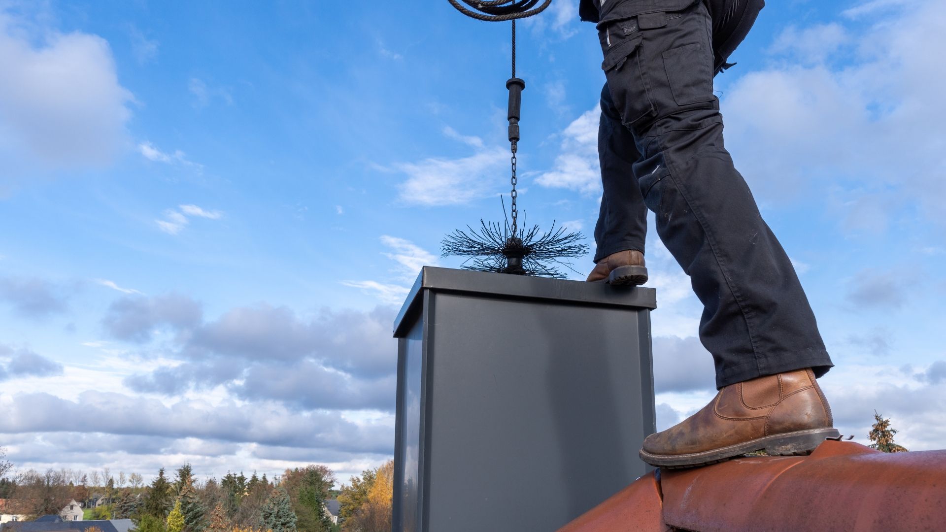 A person standing on a roof uses a rotary chimney brush to clean a black chimney flue against a bright blue sky.