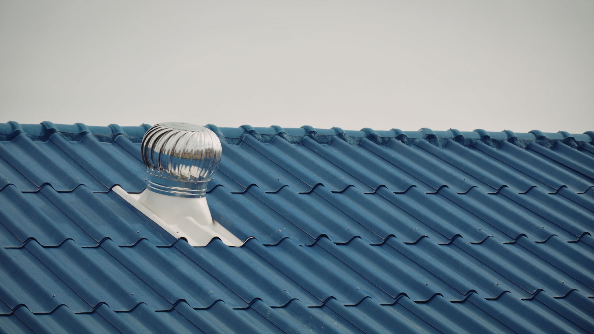 A metallic, spherical roof ventilation turbine mounted on a blue, corrugated metal roof against a grey sky.