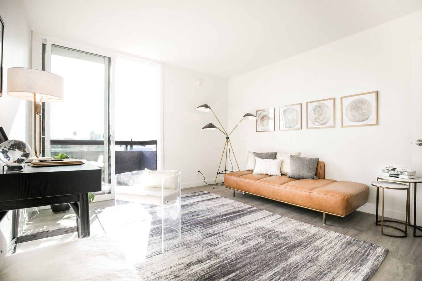 A bright, modern living room featuring a tan leather sofa, a large grey patterned rug, and a desk near a balcony window.