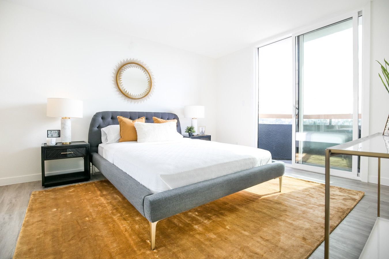 A modern bedroom with a gray tufted bed, white bedding, gold rug, a circular mirror, and sliding glass balcony doors.