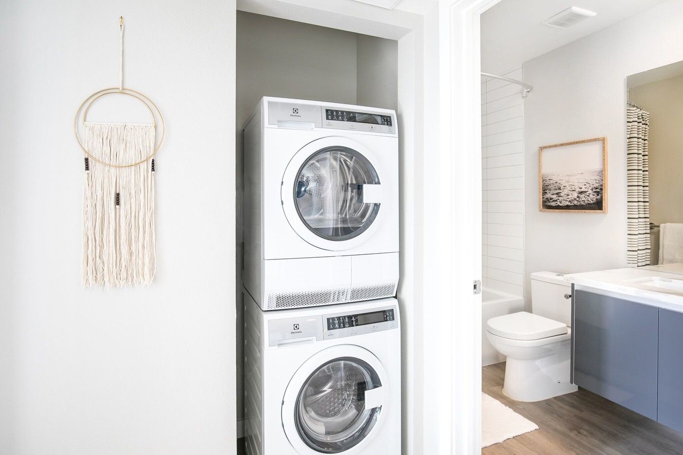 A stacked washer and dryer in a small closet, adjacent to a brightly lit bathroom with a toilet and vanity.