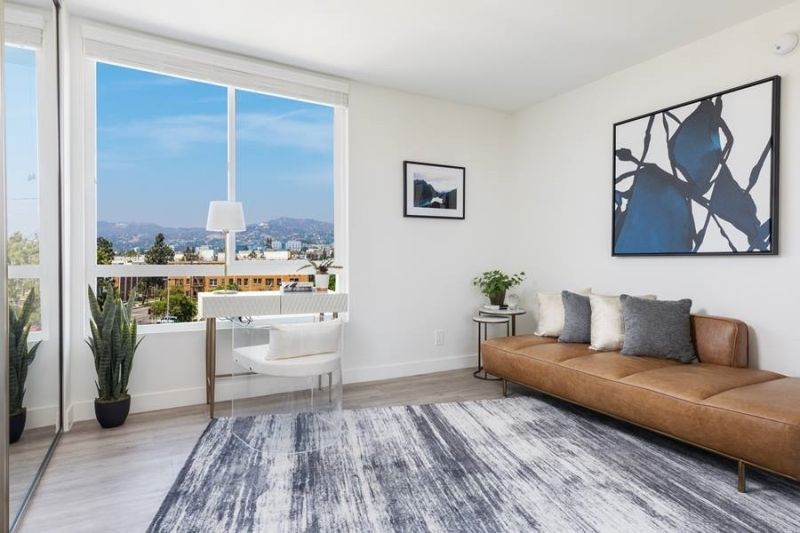 A bright room with a brown leather sofa, patterned rug, desk by a large window with a view, and abstract wall art.