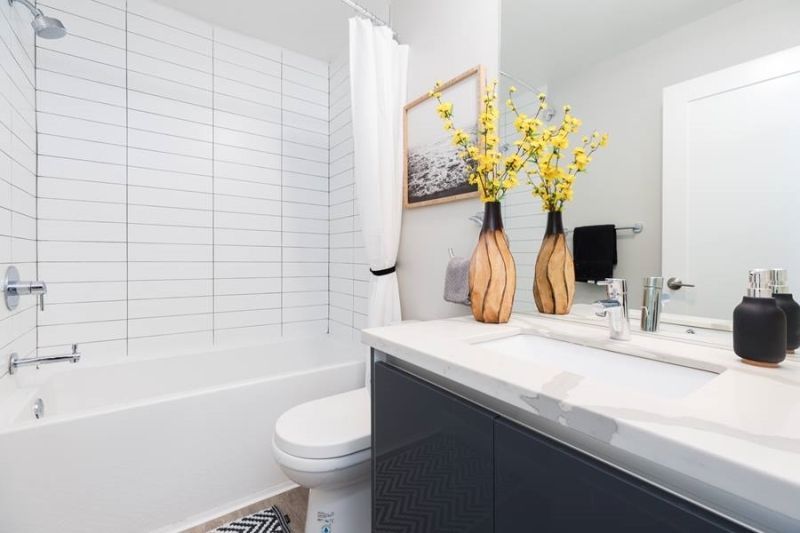 Modern bathroom with a white tiled tub, dark vanity, white countertop, and two vases of yellow flowers.