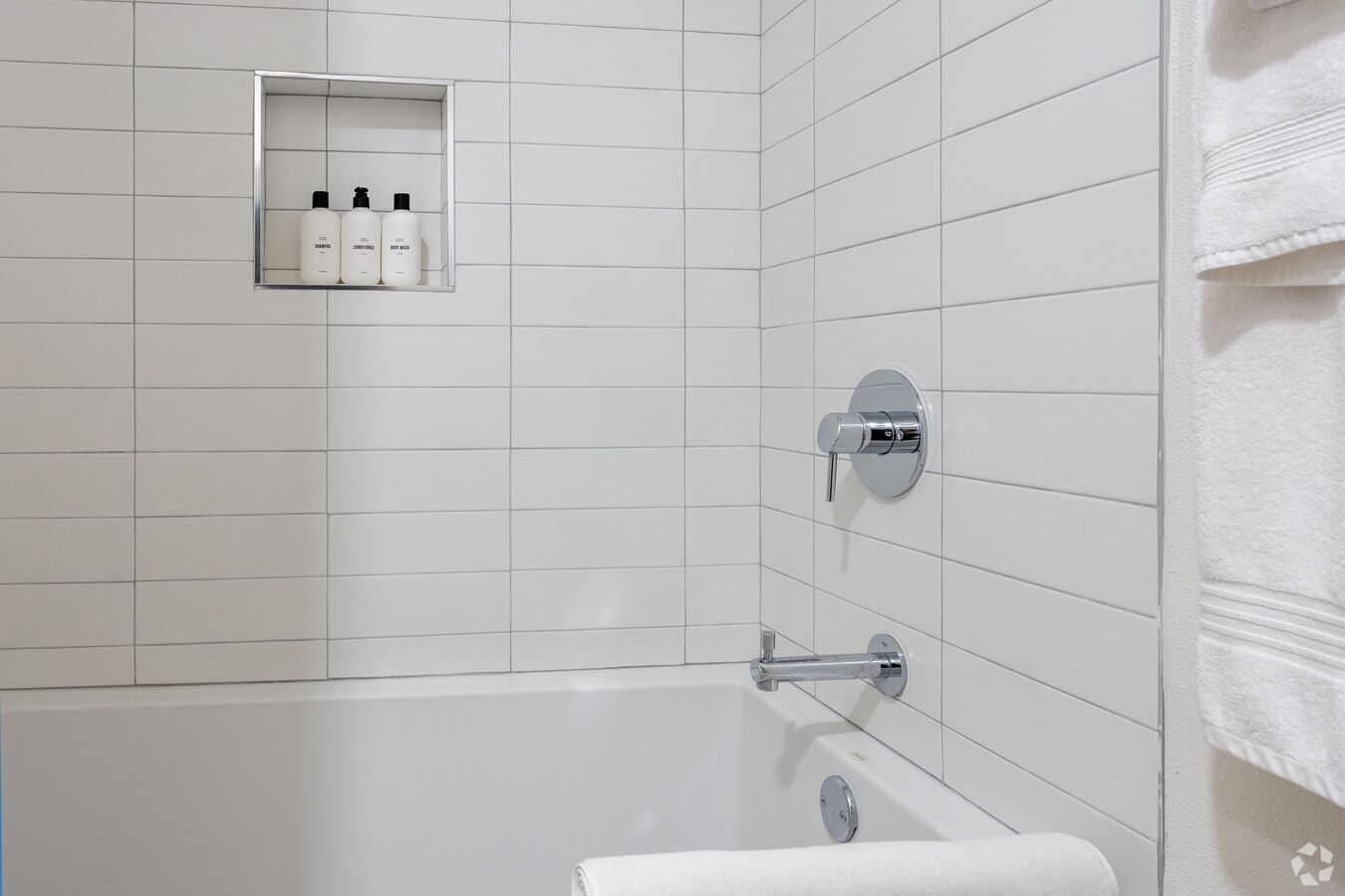 White tiled shower and bathtub with chrome fixtures, a recessed niche with toiletries, and hanging white towels.