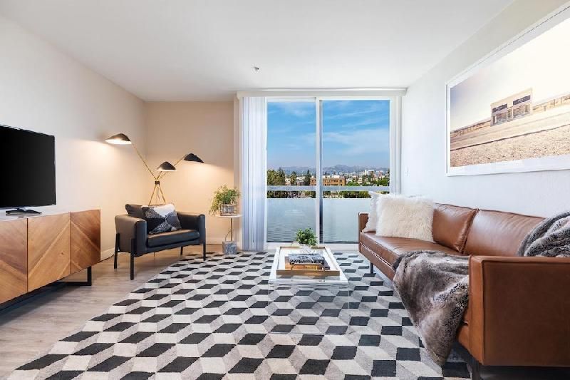 A modern living room with a geometric rug, brown leather sofa, black armchair, wood media console, and a balcony view.