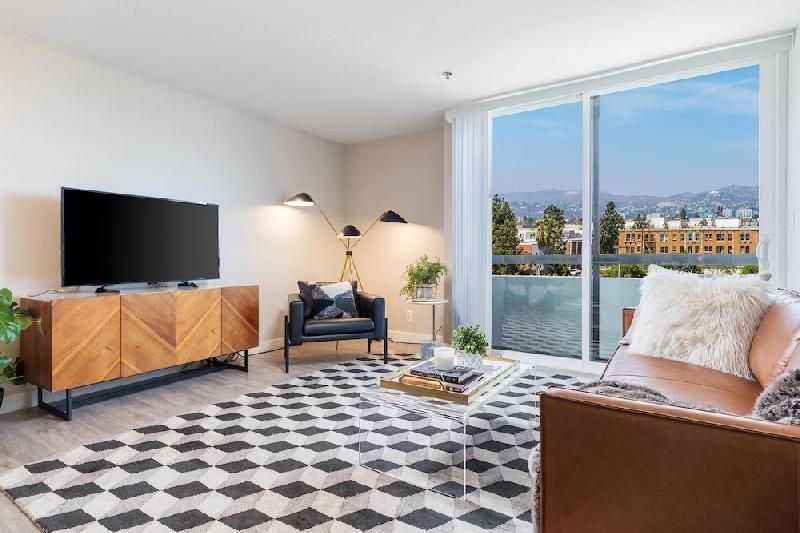 A bright living room with a geometric rug, a leather sofa, a wooden media console, and a balcony with a scenic view.