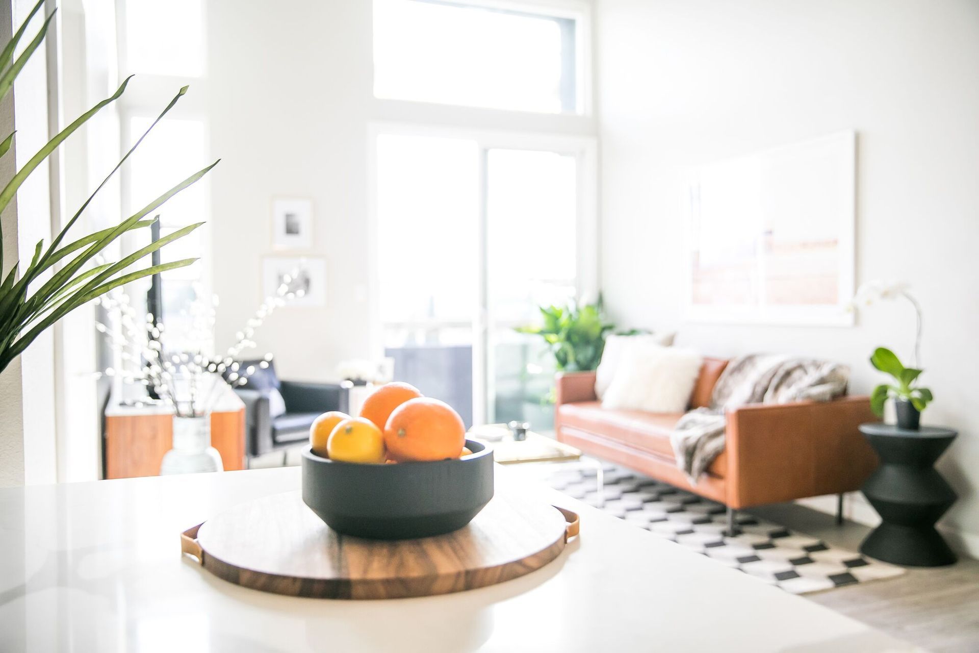A bowl of oranges on a wooden tray sits on a counter, with a blurred living room featuring a leather sofa in the background.
