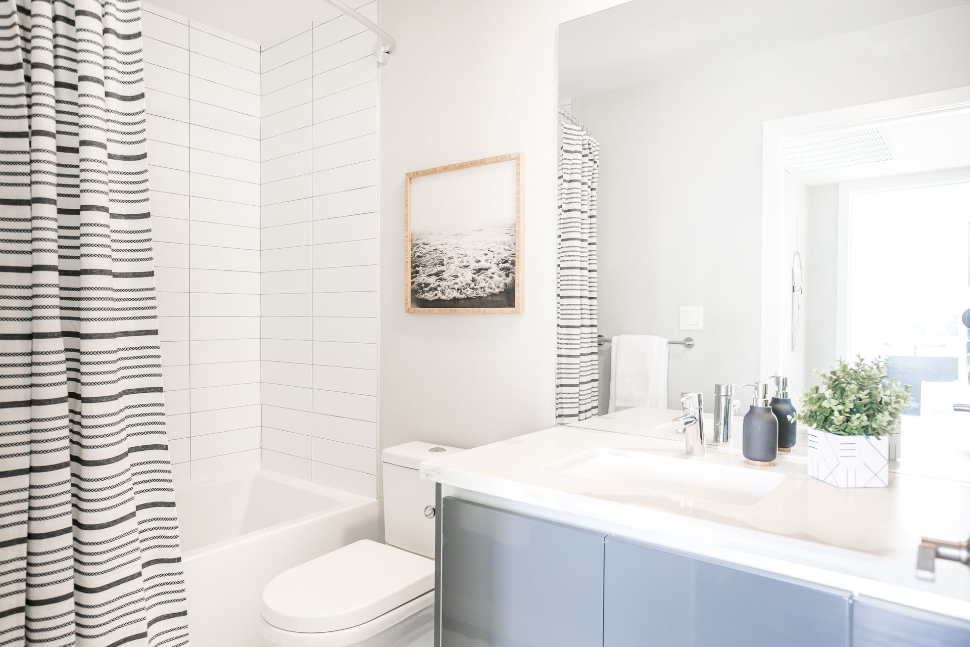 A modern bathroom featuring a white bathtub, a striped shower curtain, a grey vanity, a toilet, and a framed wall art.