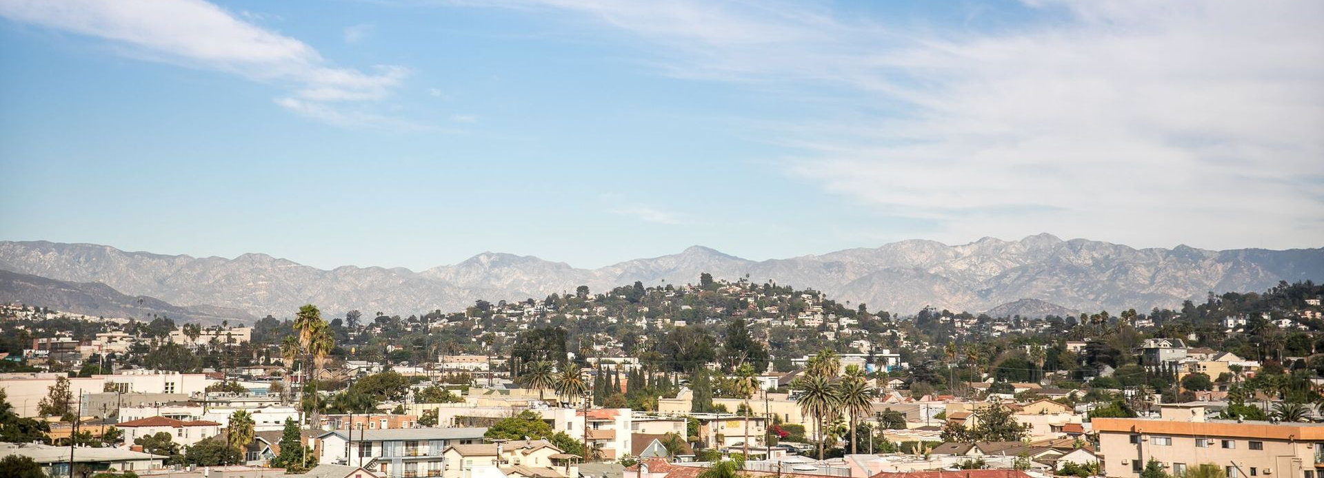 A sprawling neighborhood of homes and trees nestled beneath a range of hazy, light-colored mountains under a blue sky.